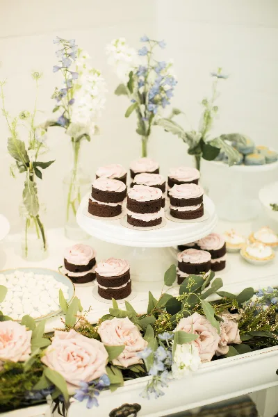 A wedding dessert display featuring layered chocolate cakes with pink frosting arranged on a white cake stand, surrounded by fresh flowers and greenery.