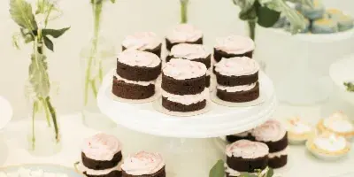 A wedding dessert display featuring layered chocolate cakes with pink frosting arranged on a white cake stand, surrounded by fresh flowers and greenery.