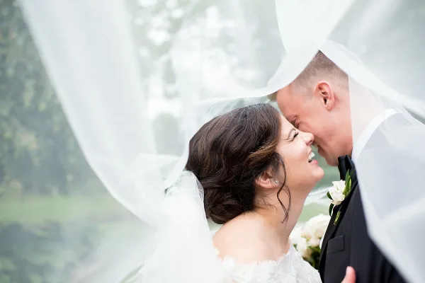Bride and groom smiling together under a flowing white veil with a boutonniere and bouquet.