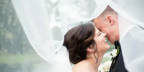 Bride and groom smiling together under a flowing white veil with a boutonniere and bouquet.