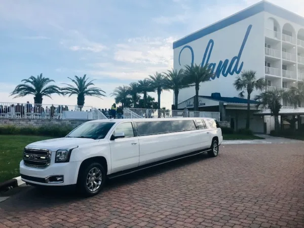 A white stretch limousine parked on a brick driveway in front of a coastal resort building with palm trees.