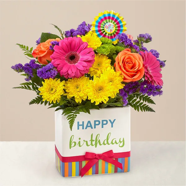 A colorful floral arrangement featuring pink gerbera daisies, yellow chrysanthemums, and orange roses in a white box with a 'Happy Birthday' message.