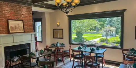 Interior view of the Claflin House dining room featuring a brick fireplace, wooden tables, and a large window overlooking a garden.