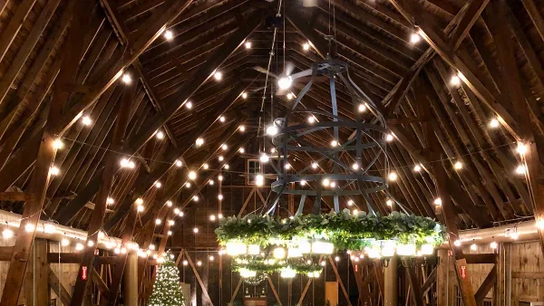Interior view of a rustic barn venue featuring exposed wooden rafters, string lights, and a large decorative chandelier.