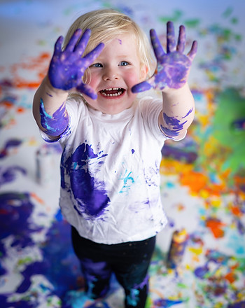 A young child with paint-covered hands and shirt smiling during a studio art photography session.