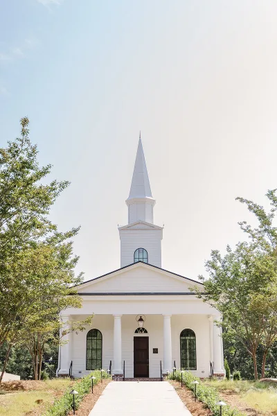 Exterior view of a white wedding chapel with a tall steeple, surrounded by trees and a paved walkway.