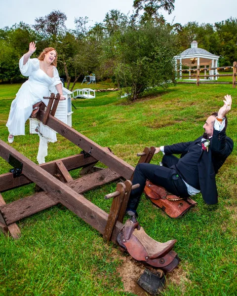 A bride in a white dress and a groom in a black suit playfully posing on a wooden seesaw structure in a grassy outdoor venue.