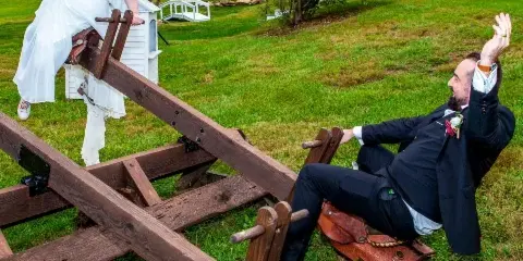 A bride in a white dress and a groom in a black suit playfully posing on a wooden seesaw structure in a grassy outdoor venue.