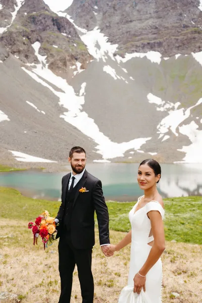 A bride and groom holding hands in front of a blue alpine lake with snow-capped mountains in the background.