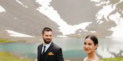 A bride and groom holding hands in front of a blue alpine lake with snow-capped mountains in the background.