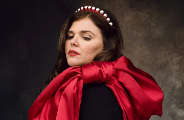 A professional studio portrait of a woman with dark hair wearing a pearl headband and a red satin scarf against a dark textured background.