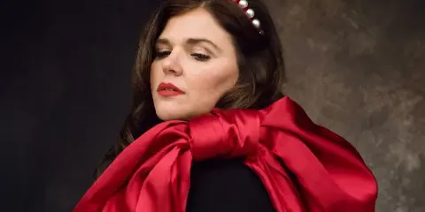 A professional studio portrait of a woman with dark hair wearing a pearl headband and a red satin scarf against a dark textured background.