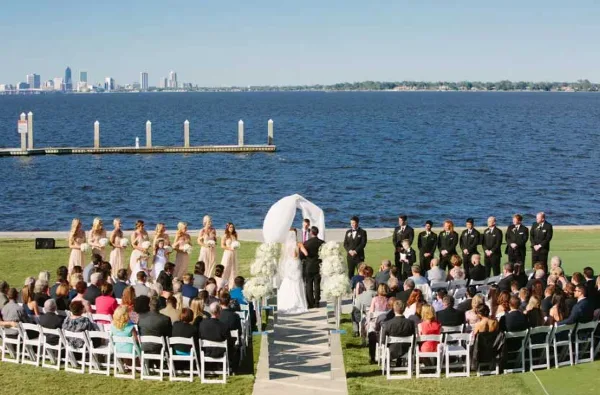 A wedding ceremony taking place on a lawn overlooking a river with a city skyline in the background.
