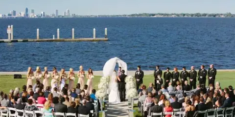 A wedding ceremony taking place on a lawn overlooking a river with a city skyline in the background.
