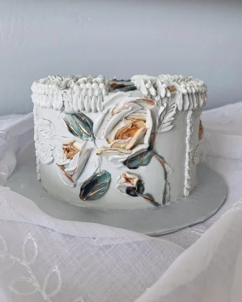 A white designer cake decorated with sculpted white flowers, teal leaves, and gold accents, sitting on a silver cake board.