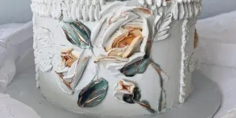A white designer cake decorated with sculpted white flowers, teal leaves, and gold accents, sitting on a silver cake board.