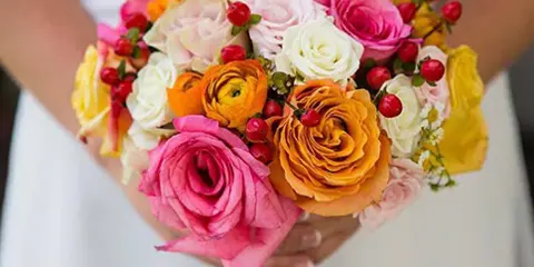 A colorful wedding bouquet held by a bride, featuring pink and orange roses, white ranunculus, and red berries.