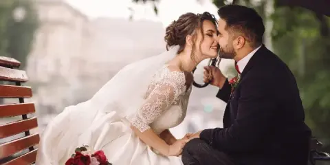 Bride and groom sharing a moment on a bench with a bouquet of red and pink roses