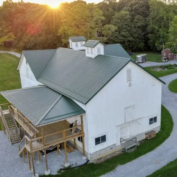 Aerial view of a large white barn with a green metal roof and wooden deck, set in a rural landscape with trees and a gravel driveway.