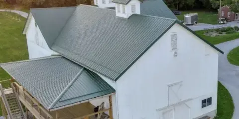 Aerial view of a large white barn with a green metal roof and wooden deck, set in a rural landscape with trees and a gravel driveway.
