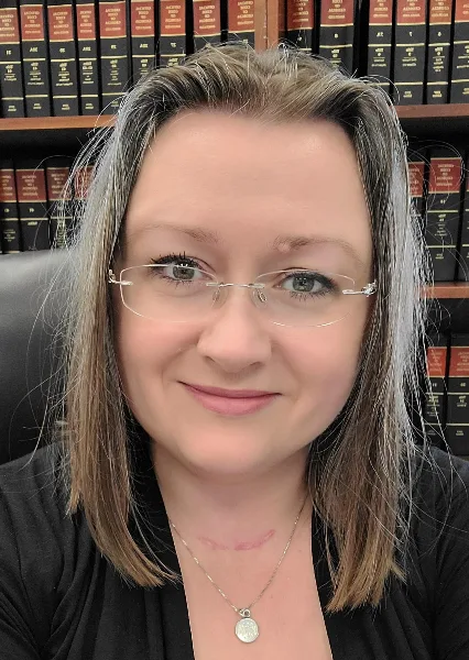 Portrait of a woman with glasses and a necklace sitting in front of a bookshelf of legal volumes.