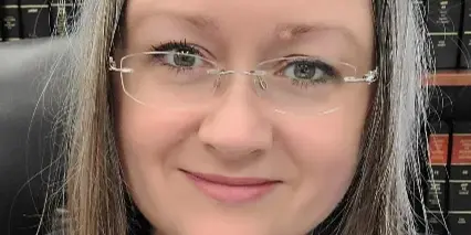 Portrait of a woman with glasses and a necklace sitting in front of a bookshelf of legal volumes.