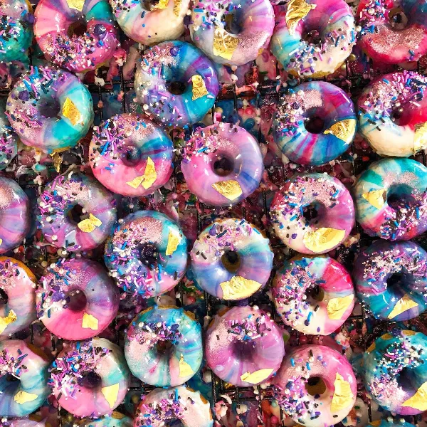 A close-up view of numerous donuts with vibrant pink, blue, and purple swirl glazes, topped with colorful sprinkles and gold leaf flakes.