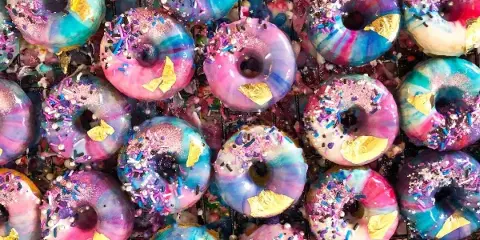 A close-up view of numerous donuts with vibrant pink, blue, and purple swirl glazes, topped with colorful sprinkles and gold leaf flakes.