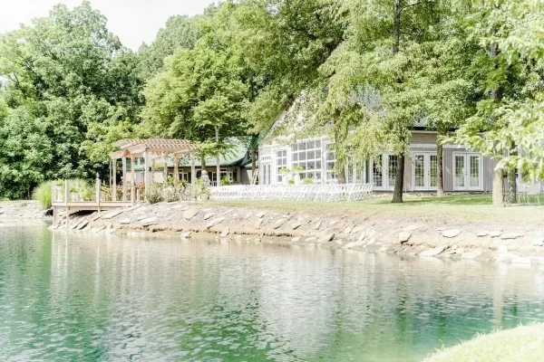 A wide shot of a wedding venue featuring a large building with a reception hall, a wooden pergola, and a pond in the foreground.