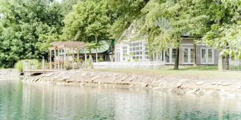 A wide shot of a wedding venue featuring a large building with a reception hall, a wooden pergola, and a pond in the foreground.