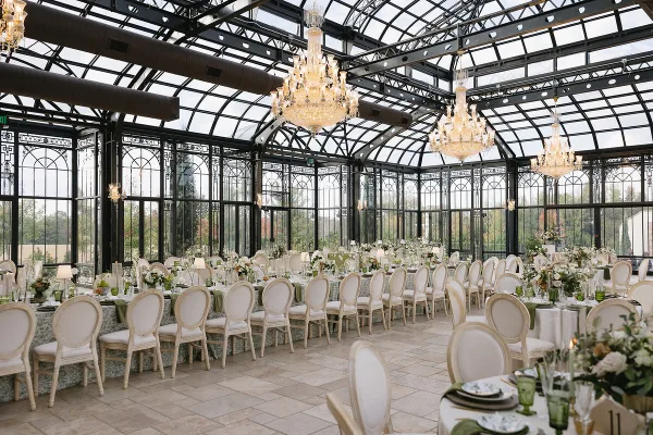 Luxury wedding reception setup in a glass conservatory with white chairs, floral centerpieces, and chandeliers.