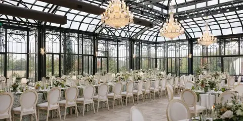 Luxury wedding reception setup in a glass conservatory with white chairs, floral centerpieces, and chandeliers.