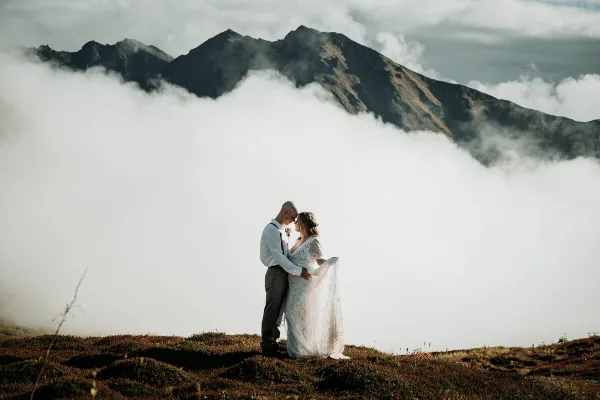 A bride and groom standing on a grassy ridge with a mountain peak and clouds in the background.