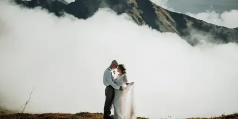 A bride and groom standing on a grassy ridge with a mountain peak and clouds in the background.