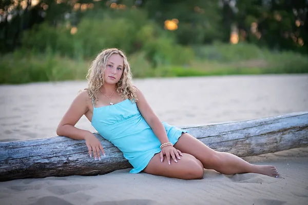 A young woman with curly blonde hair wearing a light blue dress sits on a sandy beach leaning against a large piece of driftwood.