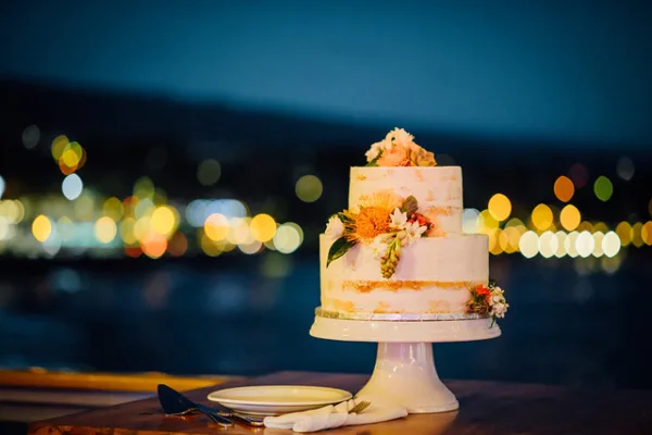 A two-tier semi-naked wedding cake decorated with fresh flowers on a white pedestal stand, set against a blurred background of city lights at night.
