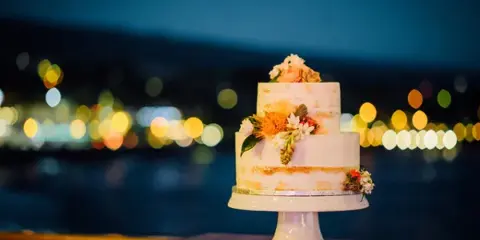 A two-tier semi-naked wedding cake decorated with fresh flowers on a white pedestal stand, set against a blurred background of city lights at night.