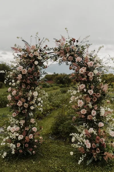 A large outdoor wedding archway decorated with lush greenery, dusty pink roses, and white flowers.