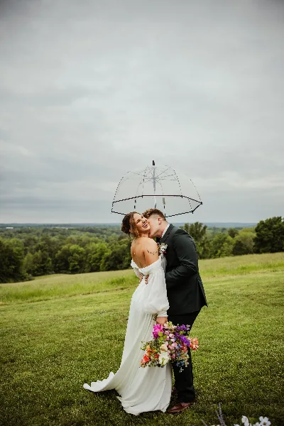 A bride and groom kissing under a clear umbrella in a grassy field.
