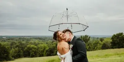A bride and groom kissing under a clear umbrella in a grassy field.