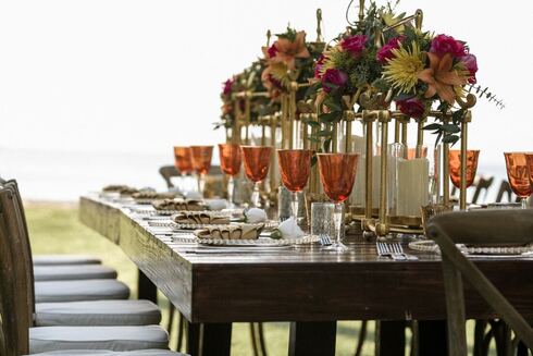 A long wooden table set for a catering event with plated food, amber glassware, and floral centerpieces.