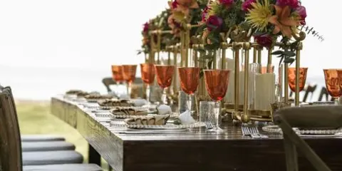 A long wooden table set for a catering event with plated food, amber glassware, and floral centerpieces.