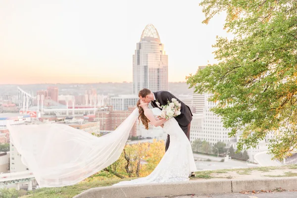A bride and groom sharing a kiss on a hilltop with the Cincinnati city skyline in the background.