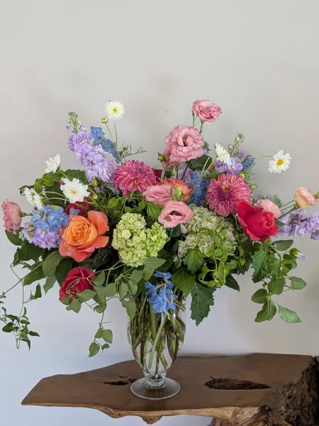 A colorful floral arrangement featuring roses, dahlias, and hydrangeas in a glass vase on a wooden table.