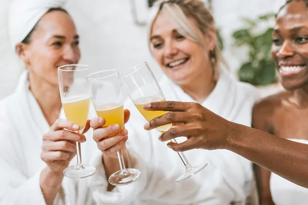Three women in white spa robes smiling and toasting with champagne flutes.