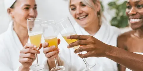 Three women in white spa robes smiling and toasting with champagne flutes.