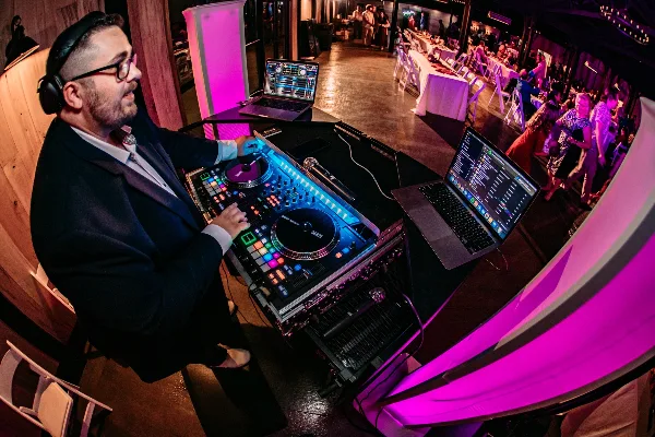 A DJ in a suit mixing on a Pioneer controller at a wedding reception with guests dancing under pink lighting.