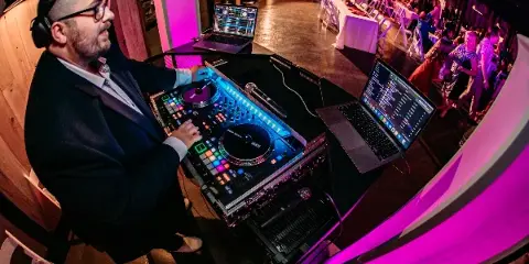 A DJ in a suit mixing on a Pioneer controller at a wedding reception with guests dancing under pink lighting.