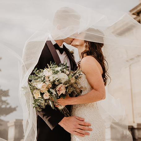 A bride and groom kissing under a veil, with the bride holding a large bouquet of white and peach flowers.