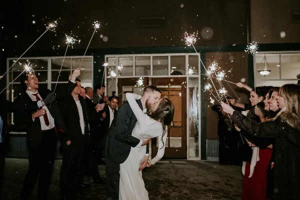 A bride and groom kiss during a sparkler send-off at night while surrounded by guests.